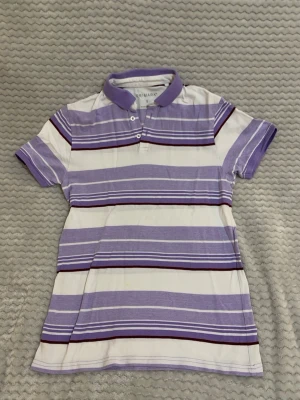 Lila och vit randig pikétröja Primark - Säljer en pikétröja från Primark i storlek S med breda lila och vita ränder samt tunna vinröda detaljer. Tröjan har klassisk krage, två knappar framtill och korta ärmar. Perfekt för en avslappnad och stilren look.