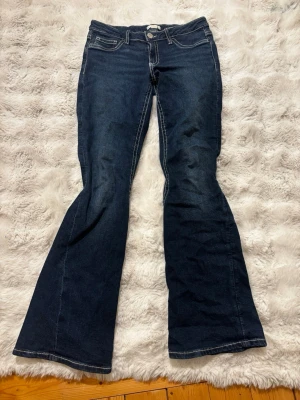 Mörkblå bootcut jeans - Säljer ett par mörkblå bootcut jeans från Gina Tricot med snygga kontrastsömmar och dekorativa bakfickor med knappar. Jeansen har låg midja och klassisk femficksdesign. Perfekta för dig som gillar en retro och avslappnad stil.