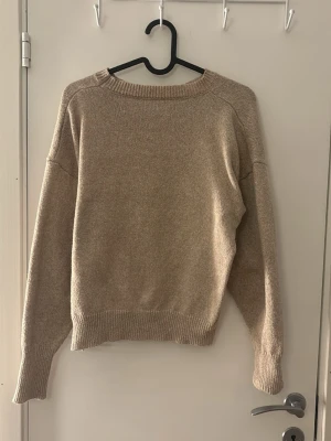 Beige stickad tröja med rund hals - En enkel och stilren beige stickad tröja med rund hals och långa ärmar. Tröjan har ribbade muddar vid ärmslut och nederkant, vilket ger en snygg och bekväm passform. Perfekt att matcha med jeans eller kjol.