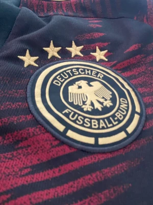 Tysklands matchtröja Adidas röd/svart - Säljer en officiell Tyskland fotbollströja från Adidas i rött och svart med guldiga detaljer och fyra stjärnor ovanför klubbmärket. Tröjan har korta ärmar, rund hals och är tillverkad i ett lätt, andningsbart material perfekt för träning eller match.
