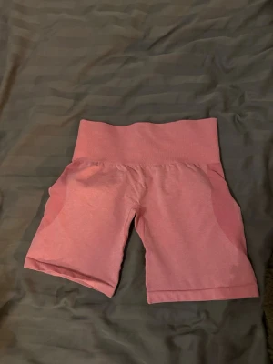 Rosa träningstights-shorts från NVGTN - Säljer ett par rosa seamless träningstights-shorts från NVGTN med hög midja och diskret logga framtill. Shortsen har en tight passform och är perfekta för gymmet eller andra träningspass. Materialet är stretchigt och bekvämt. Storlek S