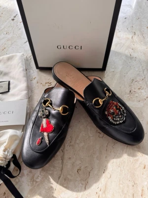Svarta Gucci loafers med brodyr - Svarta loafers från Gucci i äkta läder med guldfärgade metalldetaljer. Ena skon har broderad tiger och den andra ett svärd med hjärta och droppar. Slip-on modell med öppen häl och klassisk siluett. Snygga detaljer som sticker ut.