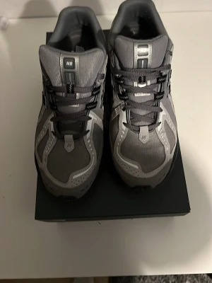 Grå New Balance sneakers - Snygga grå sneakers från New Balance med svarta och silverfärgade detaljer. Ovandelen är i mesh och syntetmaterial för bra andningsförmåga och komfort. Klassisk snörning och robust svart sula ger en sportig vibe. Perfekta för dig som gillar streetwear och stilren design.