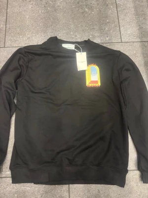  Casablanca kofta - Säljer en svart sweatshirt från Casablanca med färgglatt tryck av ett fönster och texten 'Casa Blanca' både fram och bak. Tröjan har rund hals, ribbade muddar och är gjord i mjukt bomullsmaterial. Perfekt för dig som gillar streetwear och snygga prints.
