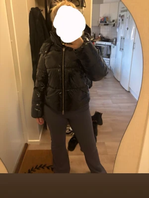 Svart glansig pufferjacka - Säljer en svart, glansig pufferjacka med hög krage och dragkedja framtill. Jackan har en boxig passform och breda kanaler som ger en riktigt puffig look. Perfekt för kalla dagar när du vill hålla värmen och ändå se stylish ut.