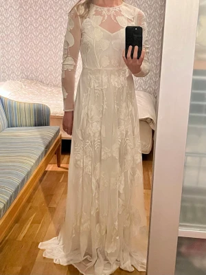 Vit långklänning med spetsdetaljer - Elegant vit långklänning med transparenta långärmar och vackra spetsdetaljer i blommigt mönster. Klänningen har rund halsringning, öppen rygg och ett flytande fall som ger en romantisk känsla. Perfekt för speciella tillfällen såsom fest eller bröllop.