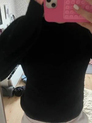 Svart stickad tröja med hög hals - Mysig svart stickad tröja med hög hals och långa ärmar. Tröjan har en enkel och stilren design som passar perfekt till många olika outfits. Materialet ser mjukt och fluffigt ut, vilket gör den extra skön att bära under kyliga dagar. Storlek S