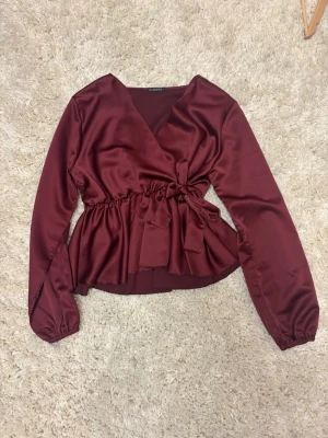 Vinröd omlottblus med knytband - Snygg vinröd omlottblus från Missäy i glansigt satintyg. Blusen har långa ärmar med resår vid handlederna och ett brett knytband i midjan som ger en markerad siluett. Perfekt för dig som gillar trendiga detaljer och en elegant look. Köpt 2024 och helt oanvänd