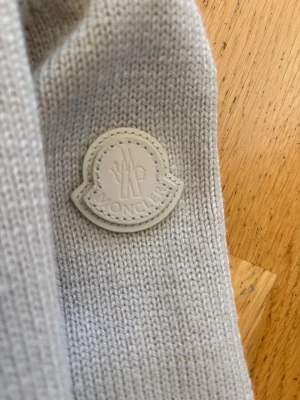 Beige dunväst från Moncler - Stilren beige dunväst från Moncler med stickade partier och quiltad front. Västen har hög krage och Moncler-logga på bröstet. Perfekt för lager-på-lager och ger en exklusiv känsla till din outfit. Materialmix av dun och stickat tyg.