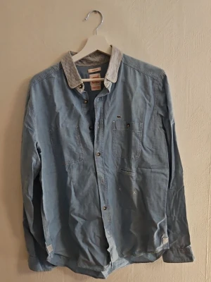 Ljusblå skjorta från Lacoste - Ljusblå skjorta från Lacoste med långärmad design och kontrasterande randig krage. Skjortan har bröstficka med liten krokodillogga och knappar framtill. Perfekt för en avslappnad men ändå stilren look.