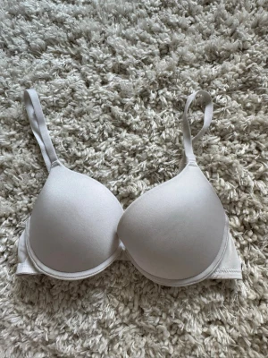 Vit vadderad t-shirt bh - En enkel och stilren vit bh med vadderade kupor, lite push up, och justerbara axelband. Bh:n har slät yta utan spets eller mönster och stängs med hakar bak. Perfekt under tighta toppar för en osynlig look. Står ej storlek men skulle uppskatta till 75A