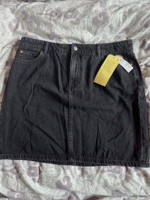 Svart jeanskjol från Lindex - Säljer en svart jeanskjol från Lindex i klassisk femficksmodell. Kjolen har knapp och dragkedja framtill samt bälteshällor i midjan. Perfekt basplagg till garderoben och enkel att styla med olika toppar.