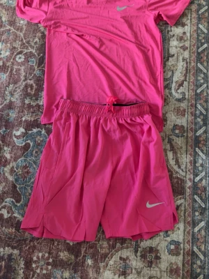 Rosa Nike miler t shirt short set - Säljer ett sportigt träningsset från Nike i stark rosa färg. Setet består av en t-shirt och ett par shorts, båda med Nike-logga. Materialet är lätt och andas, perfekt för träning eller löpning. Shortsen har resår i midjan och t-shirten har en klassisk passform.