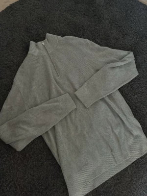 Grå stickad halvzip tröja - Säljer en stilren grå stickad tröja med halvzip och hög krage. Tröjan har ribbade muddar vid ärmslut och nederkant samt är tillverkad i ett mjukt, grovstickat material. Perfekt för lager på lager och enkel att matcha med olika outfits.