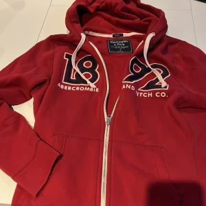 Röd huvtröja från Abercrombie & Fitch - Säljer en röd huvtröja från Abercrombie & Fitch med dragkedja och broderad logga '1892' på framsidan. Tröjan har långa ärmar och en justerbar huva med snören. Perfekt för en avslappnad stil.