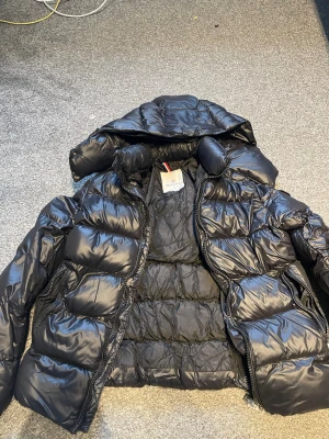 Svart dunjacka från Moncler - Snygg svart dunjacka från Moncler med glansig finish och klassisk quiltad design. Jackan har huva, dragkedja framtill och ikoniska Moncler-detaljer på insidan. Perfekt för kalla vinterdagar och riktigt varm.