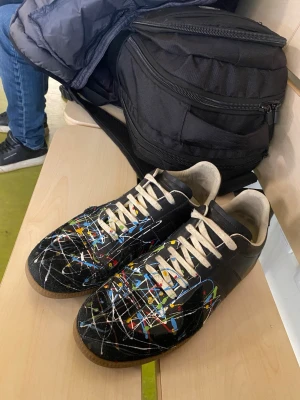 Maison Margiela med färgstänk - Unika svarta sneakers med färgstänk i blått, gult, rött och vitt. Skorna har vita snören och beige innersula. Ovandelen är i läder och sulan är i gummi. Perfekta för dig som vill sticka ut med en kreativ och konstnärlig stil.