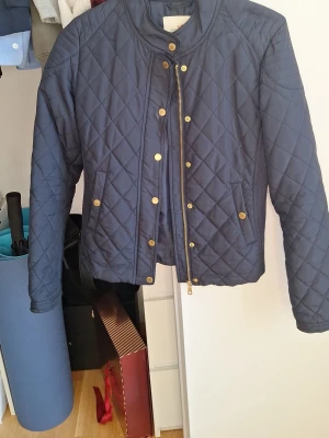 Marinblå quiltad jacka Holly & Whyte strl XS - Snygg marinblå quiltad jacka från Holly & Whyte från Lindex. Jackan har guldfärgade knappar och dragkedja, två fickor fram och en låg krage. Perfekt för dig som gillar klassisk och stilren look. Materialet är lätt vadderat och är en lätt att matcha med mycket. Bra för höst och vår