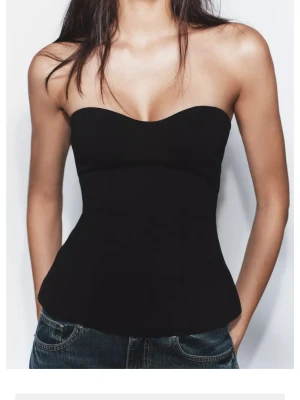 Svart axelbandslös korsettopp - Snygg svart korsettopp med axelbandslös design och lätt utsvängd nederkant. Toppen har en figurnära passform och markerad midja som ger en riktigt clean siluett. Perfekt att styla med jeans eller kjol för en trendig look.