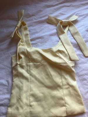 Gul topp med knytband på axlarna - Snygg ljusgul topp med breda axelband som knyts i rosetter. Modellen är figurnära med fyrkantig urringning och sömmar som ger en smickrande passform. Perfekt för sommardagar och enkel att styla till olika looks. Storlek ”S” men passar även ”M”