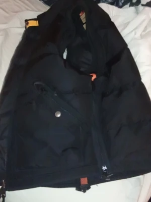 Svart Parajumpers dunväst - Svart dunväst från Parajumpers med hög krage, gul detalj vid axeln och snedställd ficka med knapp. Västen har quiltad design och dragkedja framtill. Materialet är slitstarkt och fyllningen ger bra värme. Perfekt för lager-på-lager och streetwear.