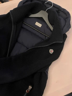 Mörkblå dunväst från Moncler - Snygg mörkblå dunväst från Moncler med stickade detaljer vid krage och sidor. Västen har dragkedja framtill, Moncler-logga på bröstet och en vadderad huva. Perfekt för lager-på-lager och riktigt stilren look.