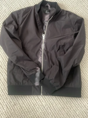 Svart bomberjacka från H&M - Stilren svart bomberjacka från H&M med klassisk ribbad krage, muddar och nederkant. Jackan har dragkedja framtill och två sidofickor. Materialet är slätt och lätt, perfekt för en trendig look. Passar till många olika outfits och är enkel att matcha. Ny och oanvänd . 