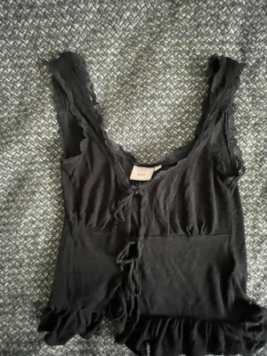 Svart spetstopp från Nelly - Säljer en svart topp från Nelly med spetsdetaljer vid axelbanden och längs urringningen. Modellen har knyt framtill och volangkant nedtill för en extra söt look. Materialet är mjukt och stretchigt, perfekt för att känna sig både bekväm och snygg.