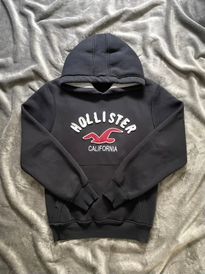 Hollister Hoodie  - Väldigt snygg Hollister Hoodie! Väldigt unik och cool design! Svart med vit textil och röd Hollister mås, storlek M-L! Ända skadan är ett litet hack (kolla sista bilden) Hör av er vid funderingar!  