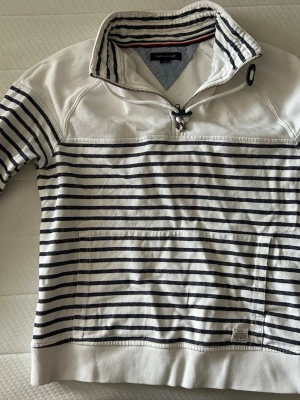Randig tröja från Tommy Hilfiger - Säljer en vit och marinblå randig tröja från Tommy Hilfiger. Tröjan har halv dragkedja, hög krage med snörning och ficka framtill. Tillverkad i mjukt bomullsmaterial. Den är riktigt schysst nu när sommaren börjar falla in. Skriv om du undrar något.🙂
