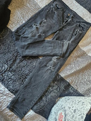 Svarta slitna jeans "Dsquared2" - Säljer ett par svarta jeans "Dsquared2" med slitningar och trasiga detaljer på flera ställen. Jeansen har rak passform, klassisk femficksmodell och knappgylf. Materialet är denim i bomull och de har en cool, avslappnad vibe.