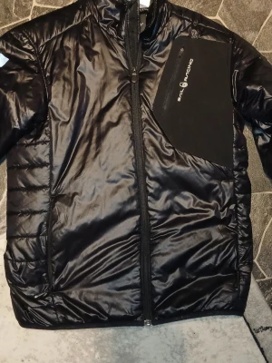 Svart pufferjacka från Sail Racing - Snygg svart pufferjacka från Sail Racing med glansig finish och dragkedja framtill. Jackan har en stor bröstficka med logga och två sidofickor. Perfekt för kalla dagar tack vare det vadderade materialet och den sportiga designen.