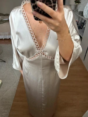 Ljus sidenklänning med djup v-ringning - Elegant ljus klänning i siden med djup v-ringning och dekorativa ringdetaljer längs urringningen och under bysten. Klänningen har vida trekvartsärmar och en mjuk, följsam passform som ger en lyxig känsla. Perfekt för dig som gillar stilrena och unika detaljer.