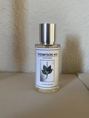 THOMPSON 41.0 parfym - THOMPSON 41.0 från One Bold Chemist är en parfym i en genomskinlig glasflaska med metalliskt silverlock. Etiketten är vit med svart text och en stilren illustration av ett blad. Flaskan har en modern och minimalistisk design.