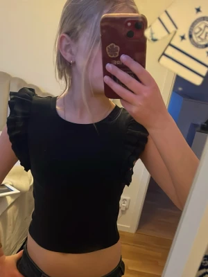 Svart topp med volangärmar - Snygg svart croppad topp med volangdetaljer på axlarna. Toppen har rund halsringning och är tillverkad i ett mjukt material som sitter bekvämt. Perfekt för dig som vill ha en stilren men ändå trendig look. Toppen är lite för liten för mig.