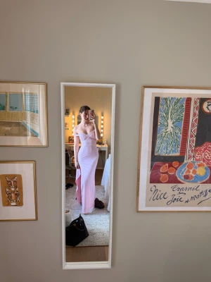 Ljusrosa långklänning offshoulder - Elegant ljusrosa långklänning med offshoulder-design och smala axelband. Klänningen har en figurnära passform och en slits framtill. Perfekt för speciella tillfällen och har fina spetsdetaljer längs urringningen.