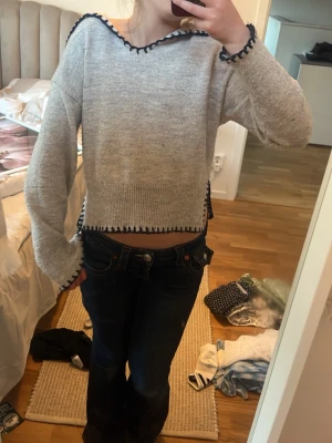 Grå stickad tröja med blå detaljer - Säljer en ljusgrå stickad tröja med mörkblå virkade kanter vid hals, ärmslut och nederkant. Tröjan har en croppad passform, vida ärmar och en avslappnad look. Perfekt för dig som gillar en trendig och unik stil. Tröjan är lite nopprig men inget som jag tycker förstör tröjan.