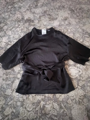 Svart omlottopp med knyt  - Stilren svart topp från mango i glansigt material med breda, halvlånga ärmar och snygg knytning i midjan. Toppen har en rak passform och en enkel rund halsringning. Perfekt för dig som gillar minimalistisk och trendig stil.