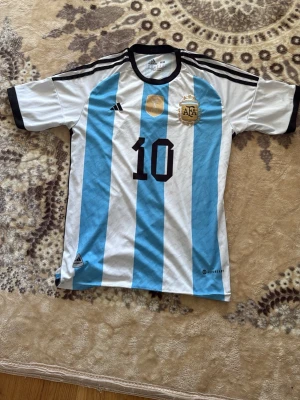 Argentina Messi matchtröja Adidas - Säljer en klassisk Argentina fotbollströja från Adidas med Messis namn och nummer 10. Tröjan har blå och vita ränder, svarta detaljer och AFA-emblem på bröstet. Perfekt för dig som älskar fotboll och vill bära landslagets färger.
