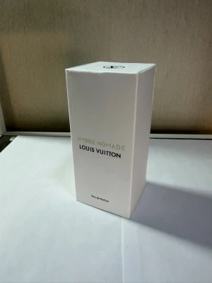 Louis Vuitton Ombre Nomade parfym - Lyxig parfym från Louis Vuitton, Ombre Nomade, i en elegant vit rektangulär kartong med diskret text i guld och svart. Förpackningen är stilren och exklusiv, perfekt för dig som gillar designerparfymer och vill ha något unikt i din samling.