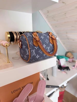  BLÅ denim handväska - Säljer en snygg blå denim handväska med klassiskt monogrammönster och detaljer i brunt läder. Väskan har två korta handtag och guldfärgade metalldetaljer. Perfekt för dig som vill ha en trendig och unik accessoar. Axelband medföljer