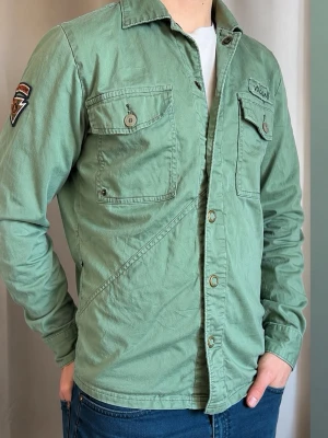 Grön overshirt från Volcom - Snygg grön overshirt från Volcom med två bröstfickor med lock och knappar, samt patch på ena ärmen. Overshirten har en avslappnad passform och är tillverkad i bomull. Perfekt som en vårjacka-lätt snygg och enkel att matcha. 
