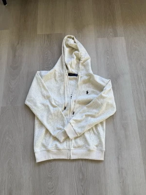 Ljusgrå hoodie från Polo Ralph Lauren - Ljusgrå hoodie med dragkedja från Polo Ralph Lauren. Klassisk design med huva och dragsko, två fickor framtill och broderad logga på bröstet. Perfekt för en avslappnad och stilren look.