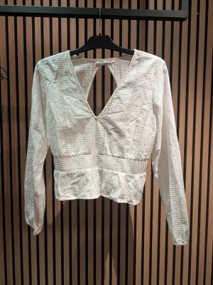 Vit spetsblus med v-ringning - Trendig vit blus i spets med djup v-ringning och snyggt hålmönster. Långärmad modell med markerad midja och öppen detalj i ryggen. Perfekt för att lyfta din outfit med en romantisk touch. Ej töjbar, liten i storleken.