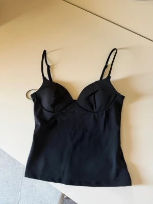 Svart body med tunna axelband - Snygg svart body med tunna, justerbara axelband och formpressade kupor. Materialet är slätt och stretchigt för en bekväm passform. Perfekt att styla under skjortor eller tröjor för en clean look.