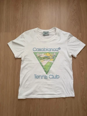 Vit Casablanca Tennis Club t-shirt - Säljer en vit t-shirt från Casablanca Tennis Club med färgglatt tryck framtill i grönt, gult och blått. Klassisk rund hals och korta ärmar. Perfekt för dig som gillar sportig och stilren design.