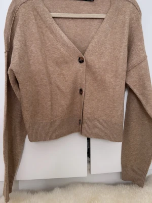 Beige kofta från Vero Moda - Snygg beige kofta från Vero Moda med v-ringning och tre knappar framtill. Koftan har långa ärmar och ribbade muddar vid ärmslut och nederkant. Perfekt att slänga över en t-shirt eller linne för en chill look.