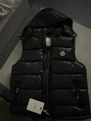 Svart dunväst från Moncler med huva - Snygg svart dunväst från Moncler med glansig finish, avtagbar huva och två dragkedjefickor. Klassisk Moncler-logga på bröstet och märkestag kvar. Perfekt för lager-på-lager och streetstyle.