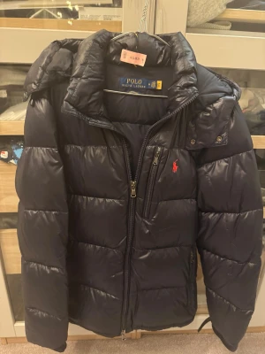 Mörkblå pufferjacka Polo Ralph Lauren - Snygg mörkblå pufferjacka från Polo Ralph Lauren med hög krage, dragkedja och två sidofickor. Jackan har det klassiska röda Polo-logot på bröstet och är tillverkad i glansigt material som håller värmen under kalla vinterdagar.