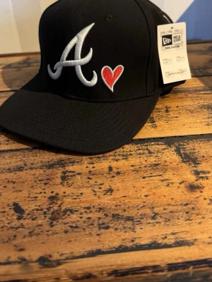 Svart New Era keps Atlanta Braves - Säljer en svart keps från New Era med Atlanta Braves-logga i vitt och ett rött hjärta framtill. På sidan finns ett broderat 1999 World Series-märke i blått, gult och rött. Klassisk rak skärm och justerbar passform. Perfekt för dig som gillar sportig streetstyle.